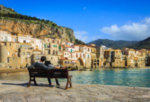 Casal sentado na frente do mar em Cefalu, na Sicília, com mar azul e casas brancas