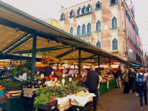 Vista do mercado de rialto, com banca de frutas, homem na frente e prédio típico ao fundo