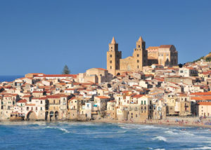 Cefalu, na Sicília, Itália, vista a partir do mar, com casas em tons claros e catedral ao fundo.