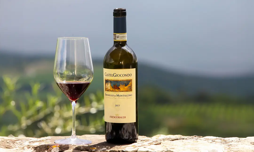 Taça de vinho tinto ao lado de garrafa de Brunello di Montalcino CastelGiocondo 2015, da vinícola Frescobaldi, com paisagem vinícola da Toscana ao fundo.