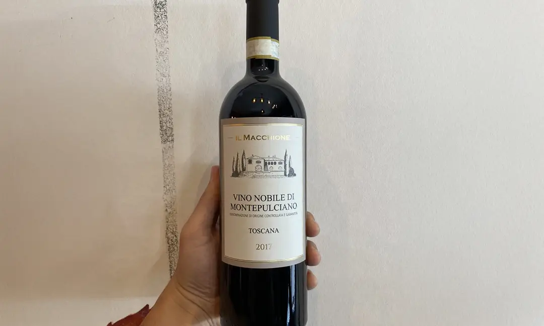 Pessoa segurando uma garrafa de vinho Il Macchione Vino Nobile di Montepulciano 2017, com rótulo elegante em destaque contra fundo artístico.