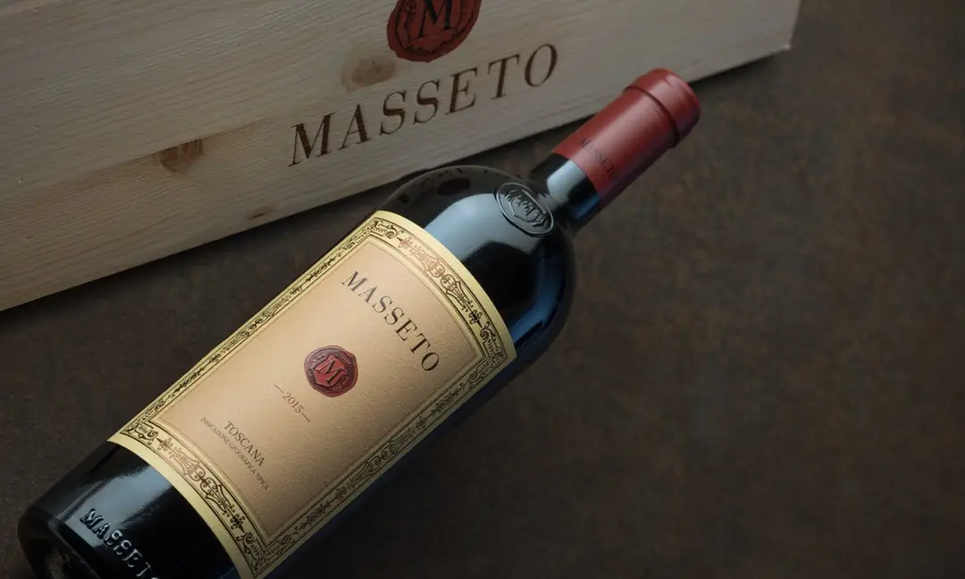 Garrafa de vinho Masseto 2015 posicionada sobre superfície escura ao lado de caixa de madeira personalizada, destacando o rótulo elegante e luxuoso do renomado vinho toscano